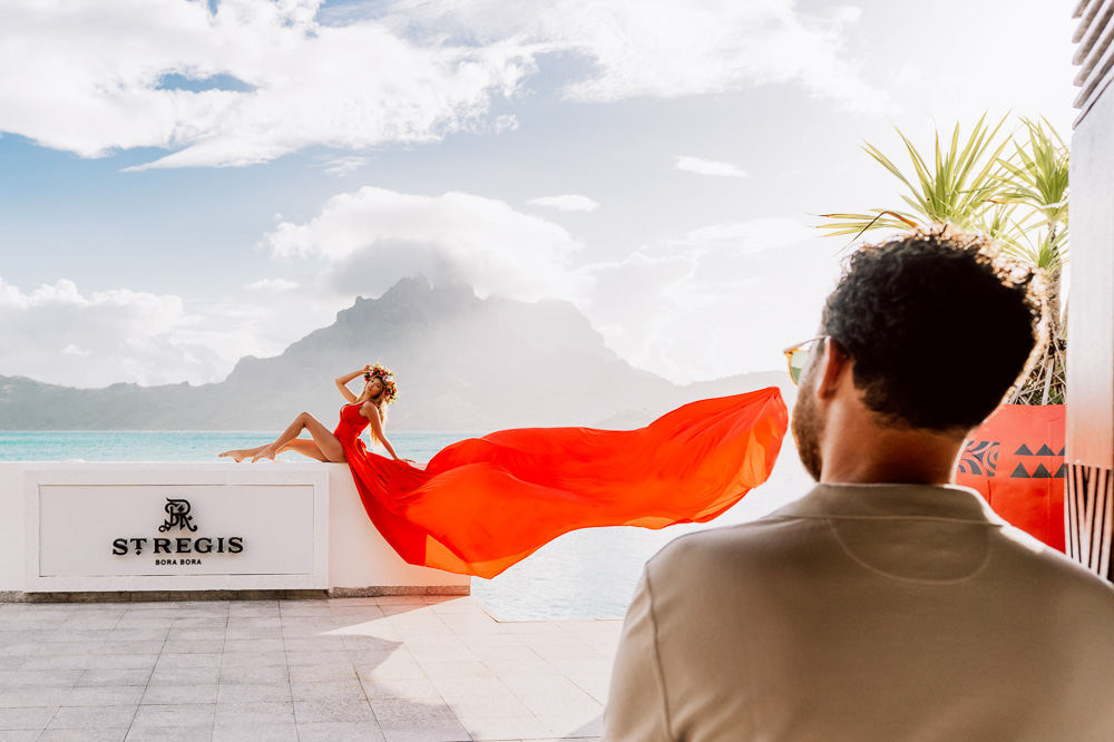 Séance photo de flying dress ou le mari regarde sa femme assise avec une longue robe rouge qui vole dans le airs